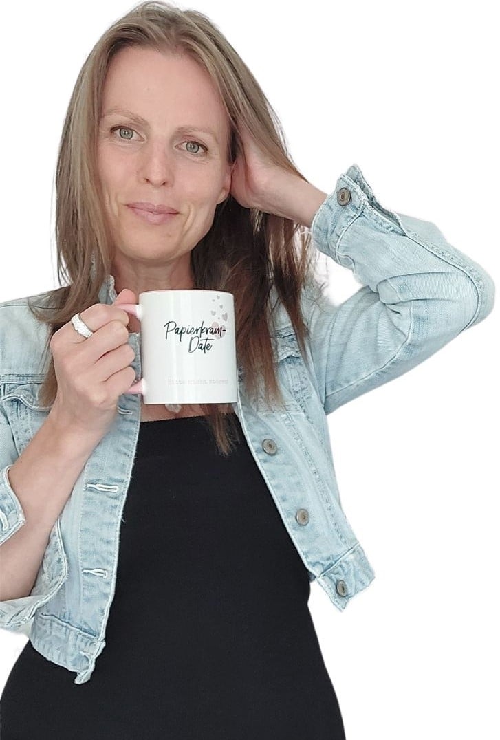 Eine Person mit langen Haaren hält einen weißen Becher in einer Hand und berührt mit der anderen ihr Haar. Sie trägt eine helle Jeansjacke über einem schwarzen Oberteil.