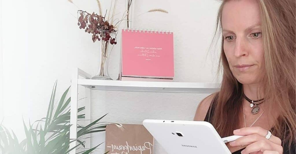 Eine Frau steht vor einem Regal mit Pflanzen und hält ein weißes Tablet in der Hand. Auf dem Regal sind ein rosa Kalender und ein Schild zu sehen.