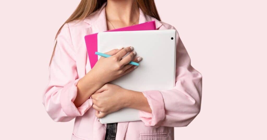 Eine Person in einer rosa Jacke hält ein weißes Notizbuch, einen lila Ordner und einen blauen Stift vor einem hellrosa Hintergrund.