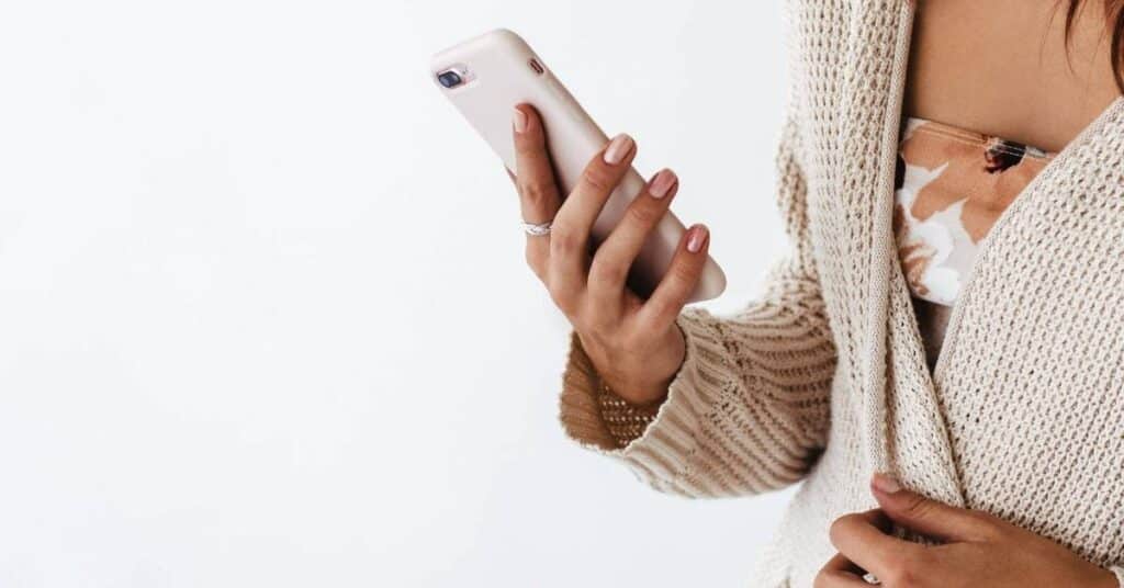 Person in einem beigen Pullover, die ein Smartphone mit einer hellrosa Hülle hält.