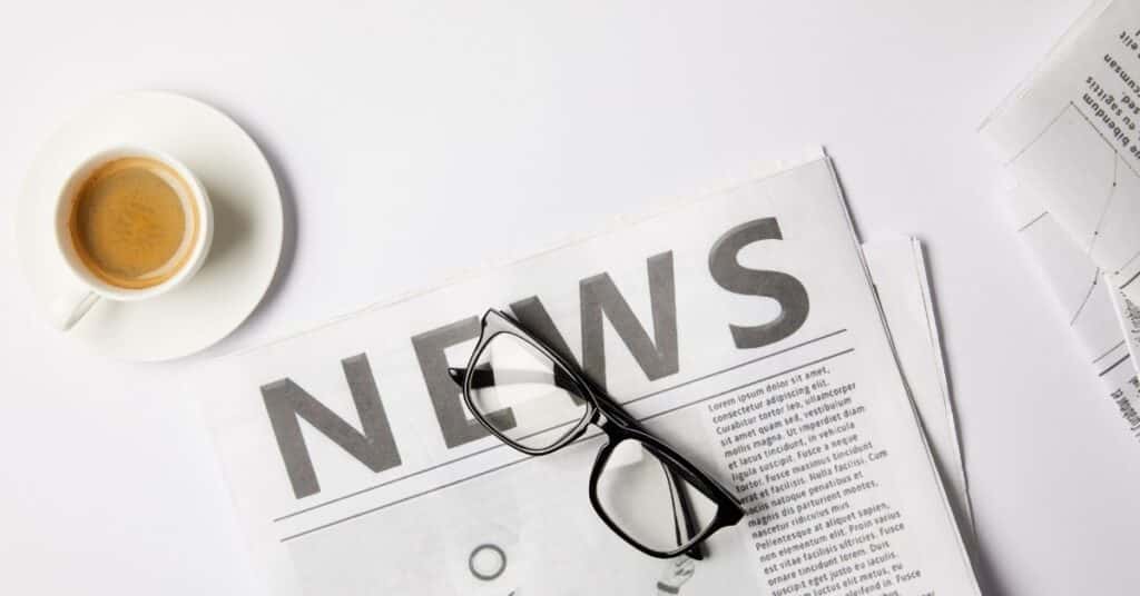 Auf einem Tisch liegt eine Zeitung mit der Schlagzeile „NEWS“, dazu eine Brille und eine Tasse Espresso.