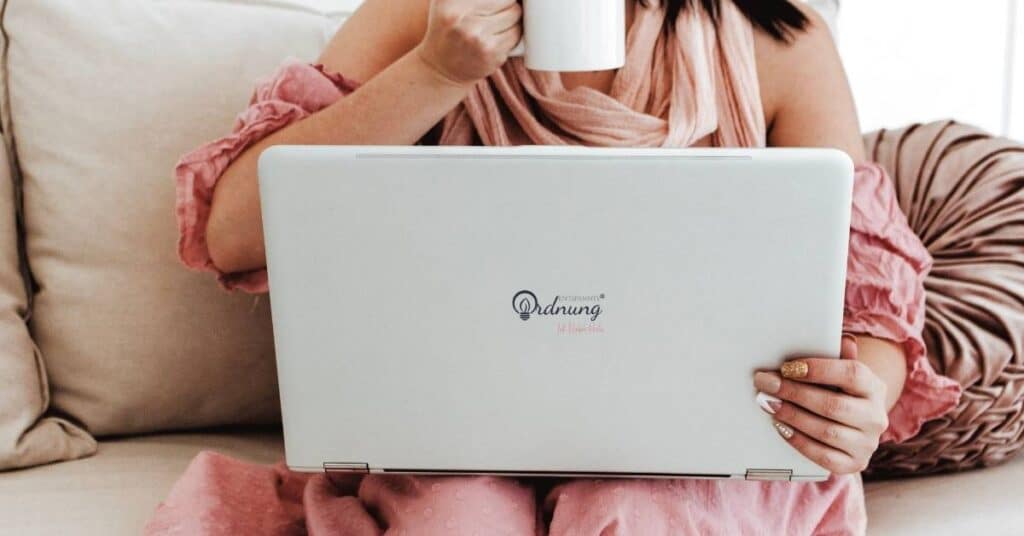 Eine Person im rosa Outfit sitzt auf einer Couch und hält mit beiden Händen eine weiße Tasse und einen Laptop.