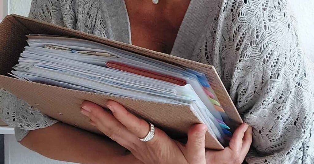 Eine Person hält einen großen beigen Ordner voller Papiere und Registerteiler und trägt einen gemusterten grauen Pullover.