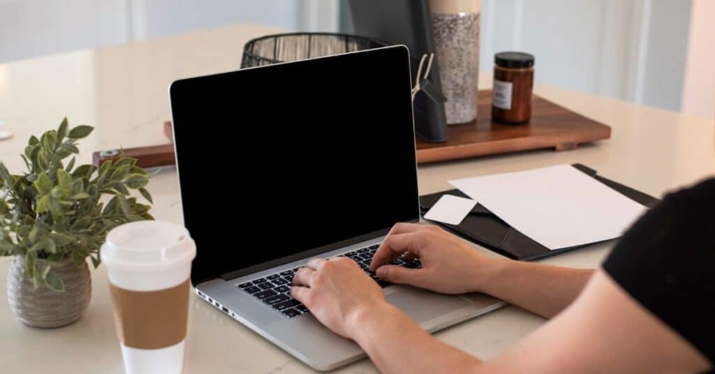 Eine Person tippt auf einem Laptop mit leerem Bildschirm. Auf dem Schreibtisch stehen eine Kaffeetasse, eine Pflanze und Büromaterial.