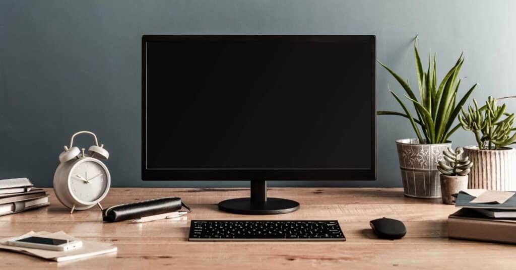 Ein aufgeräumter Schreibtisch mit Monitor, Tastatur und Maus. Daneben Wecker, Notizbücher, Smartphone, Stifteetui und Topfpflanzen.