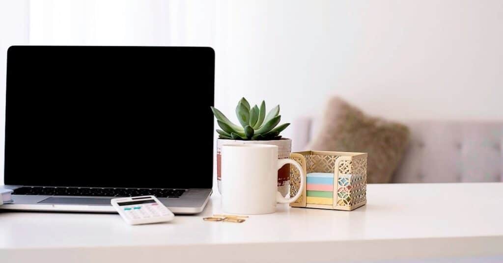Laptop auf einem weißen Schreibtisch mit einer Tasse, einer kleinen Pflanze, einem Taschenrechner, Büroklammern und einer goldenen Schachtel mit bunten Haftnotizen.