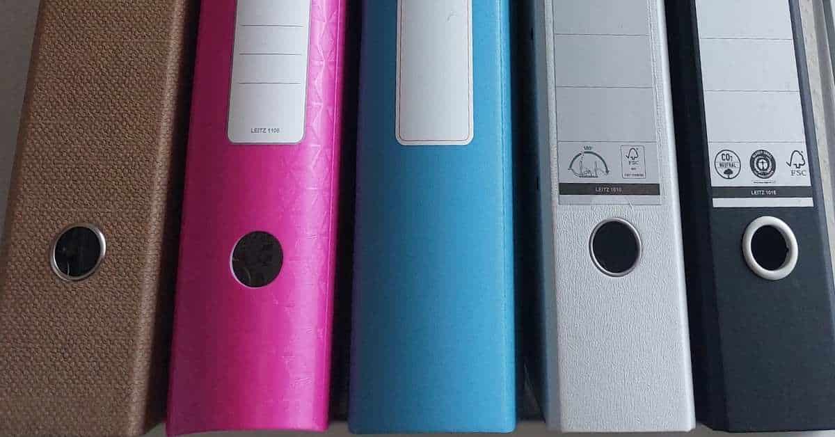 Fünf stehende Ringbücher in verschiedenen Farben: Braun, Rosa, Blau, Weiß und Schwarz, in einer Reihe angeordnet.