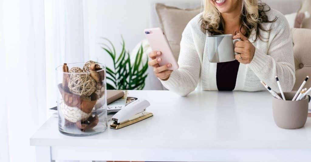 Person sitzt an einem Schreibtisch und hält eine Tasse und ein Smartphone. Auf dem Schreibtisch stehen eine Pflanze, ein Bleistifthalter und ein Glasbehälter mit Dekor.