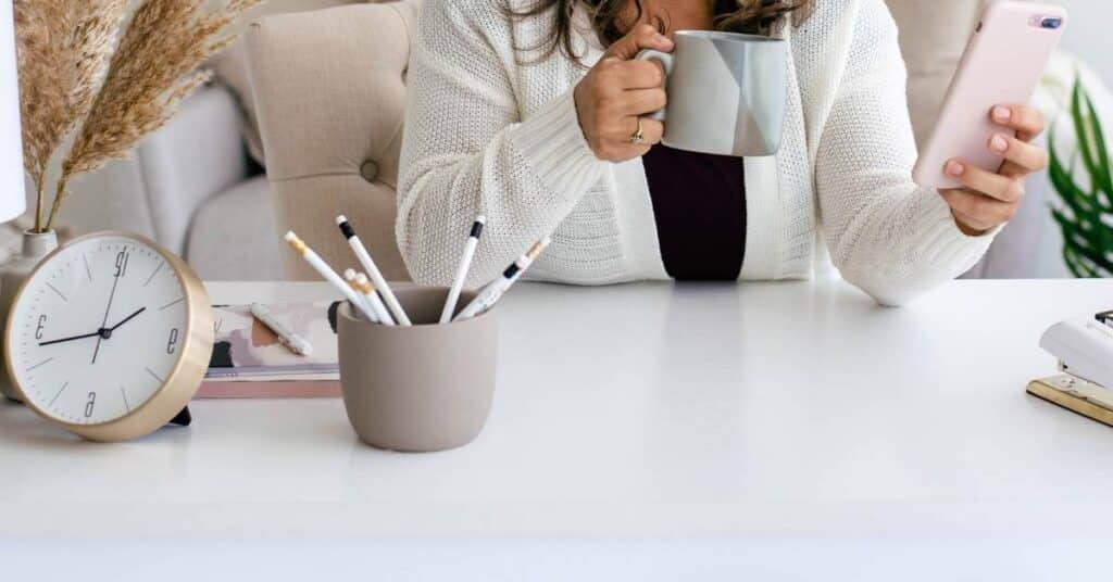 Person sitzt an einem weißen Schreibtisch und hält ein Smartphone und eine Tasse. Auf dem Schreibtisch stehen eine Uhr, eine Tasse mit Bleistiften und getrocknete Pflanzen.