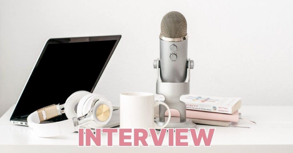 Auf einem weißen Schreibtisch liegen Mikrofon, Laptop, Kopfhörer, Tasse und Bücher. Unten prangt in großen rosa Buchstaben das Wort „Interview“.