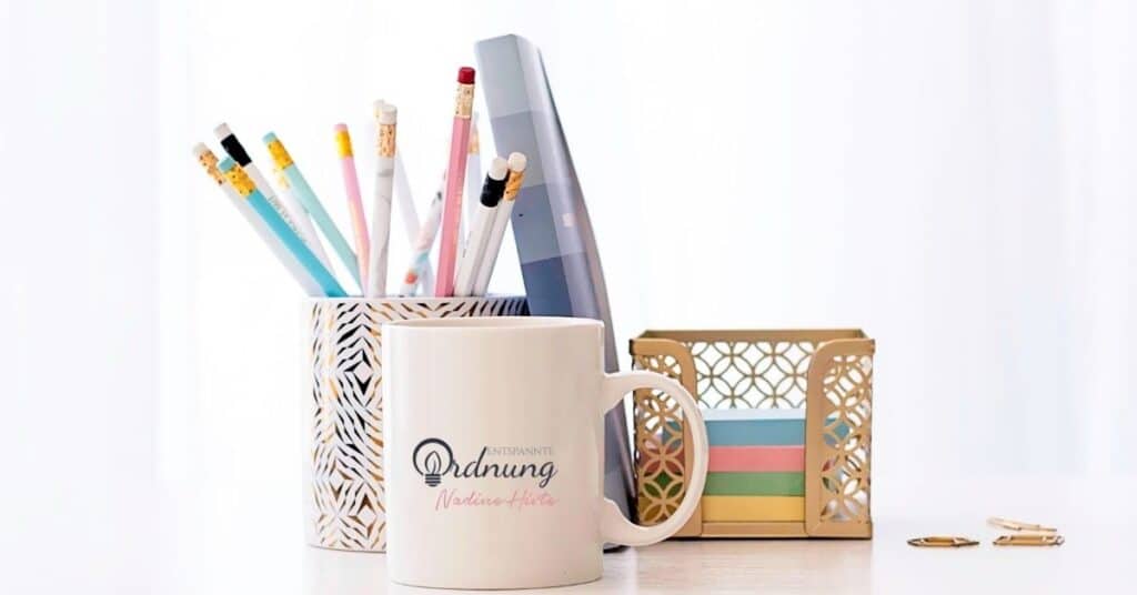 Ein Schreibtisch mit einer Tasse, einem Halter mit Bleistiften, einem Buch, einer dekorativen Box mit Haftnotizen und Büroklammern.