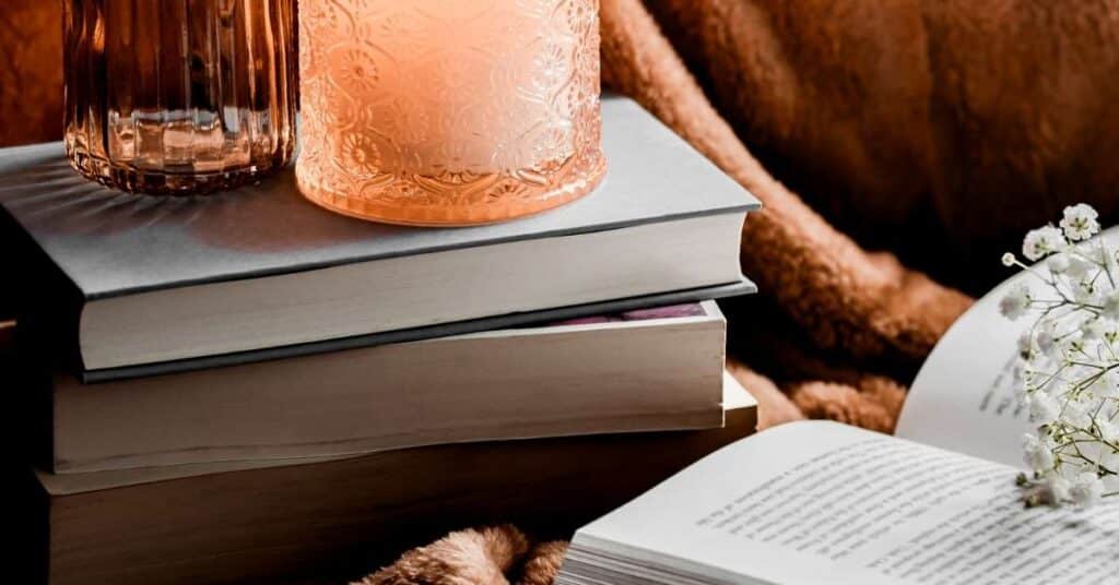 Ein Stapel Bücher mit einer brennenden Kerze darauf, ein offenes Buch daneben und eine kleine weiße Blume auf einer gemütlichen, braunen Decke.