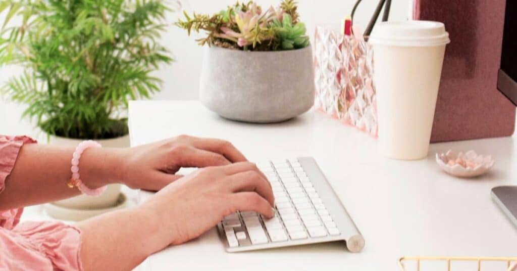 Person tippt auf einer weißen Tastatur an einem Schreibtisch mit einer Topfpflanze, einem Pappkaffeebecher und Dekorationsgegenständen in der Nähe.