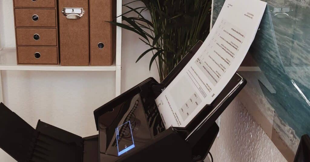Ein Dokumentenscanner scannt gerade ein Blatt Papier. In der Nähe stehen braune Aktenkartons und auf einem Regal eine Pflanze.