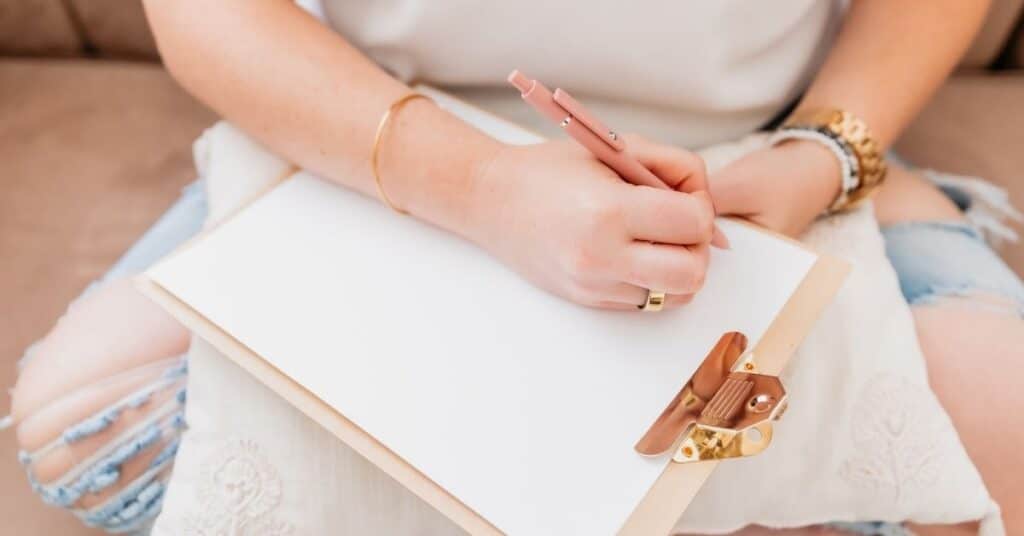 Eine Person in einem weißen Hemd und zerrissenen Jeans schreibt mit einem rosa Stift auf ein Klemmbrett.