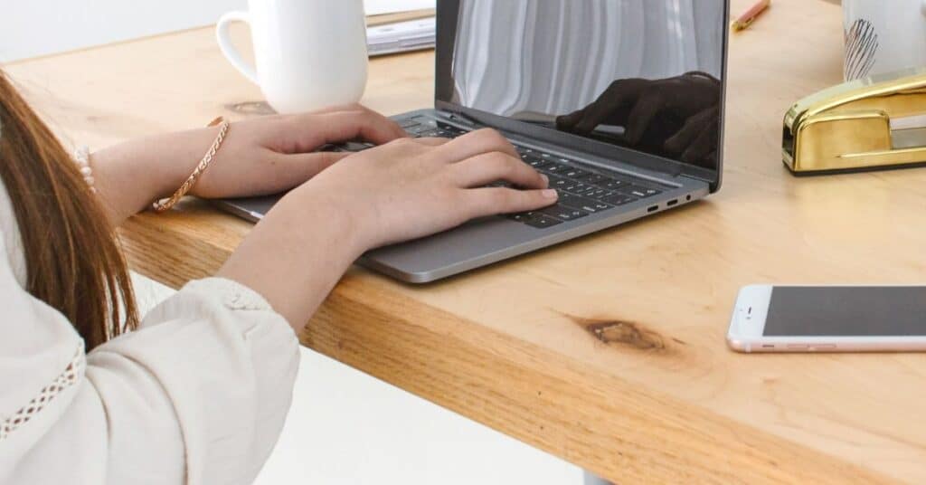 Person tippt auf einem Laptop an einem Holzschreibtisch, in dessen Nähe sich ein Telefon, eine Tasse und ein Hefter befinden.
