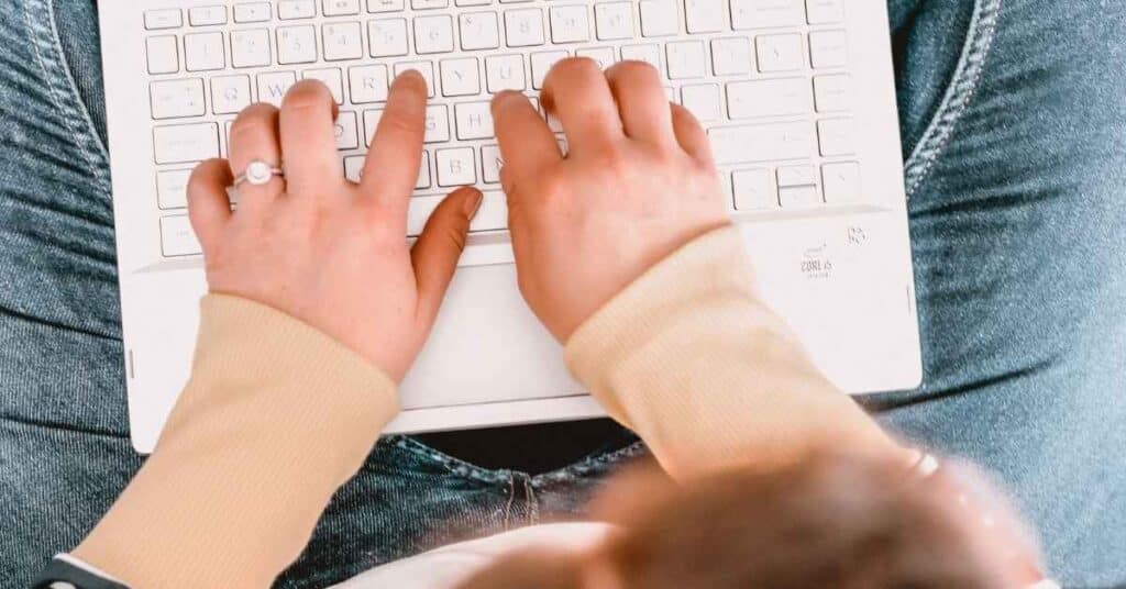 Eine Person in einem beigen Pullover tippt auf einem weißen Laptop, der auf ihrem Schoß steht.