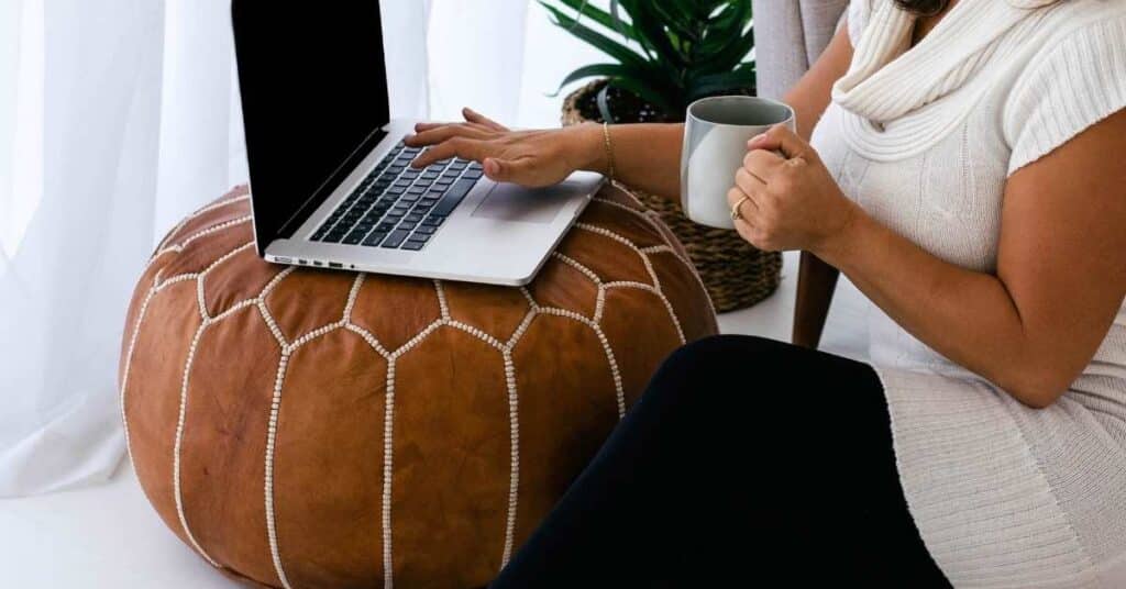 Person, die einen Laptop auf einem braunen Sitzpuff benutzt und eine Tasse hält, neben einer Pflanze in der Nähe eines Fensters.