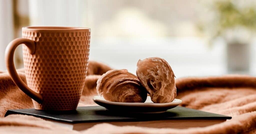 Eine strukturierte Tasse und zwei Croissants auf einem kleinen Teller stehen auf einer weichen Decke neben einem Fenster.
