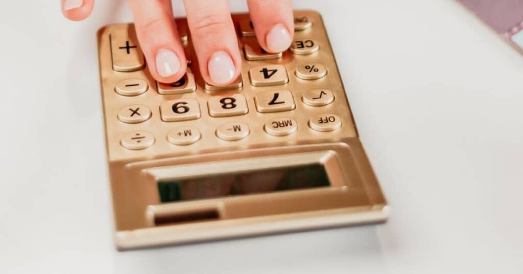 Eine Hand drückt vor einer weißen Oberfläche die Tasten eines goldenen Taschenrechners.