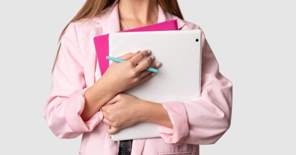 Eine Person im rosa Blazer hält Notizbücher und einen Stift vor einem hellgrauen Hintergrund.