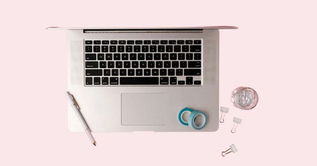 Laptop auf einer rosa Oberfläche mit einem Stift, zwei Rollen Klebeband, einer kleinen Tasse Büroklammern und Heftklammern in der Nähe.