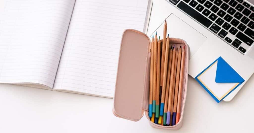 Ein aufgeschlagenes Notizbuch, ein Satz Buntstifte in einem rosa Etui, ein Laptop und ein Stapel Haftnotizen liegen auf einem Schreibtisch.