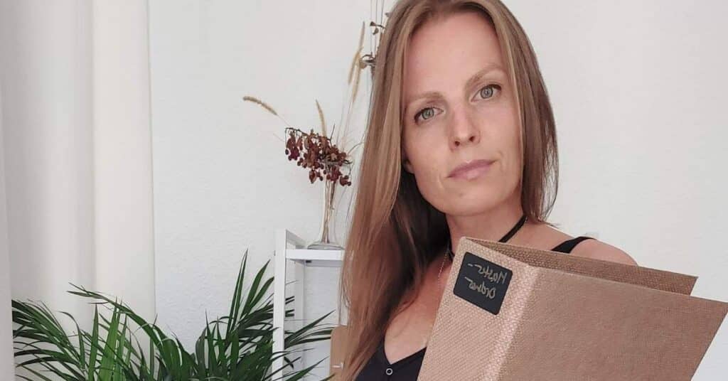 Eine Person mit langen Haaren hält ein geschlossenes Buch oder einen Ordner und steht im Innenbereich mit Pflanzen im Hintergrund.