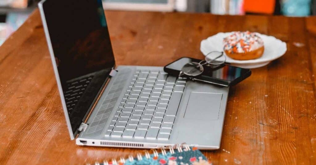 Ein Laptop auf einem Holztisch mit einer Brille, einem Smartphone, einem gemusterten Notizbuch und einem Teller mit einem Donut.