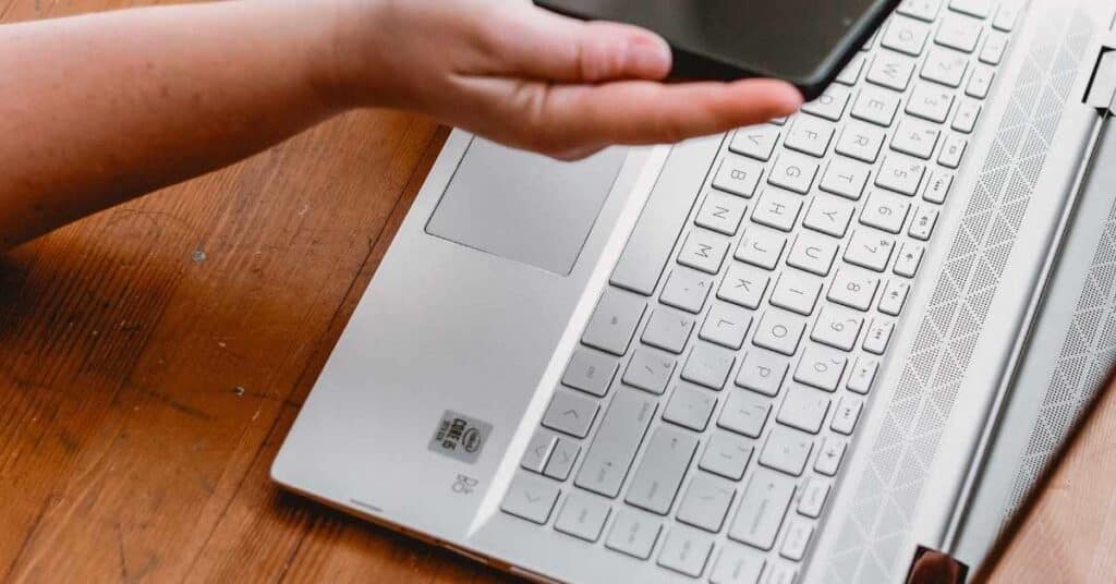 Die Hand einer Person hält ein Smartphone über einem geöffneten Laptop auf einem Holztisch.