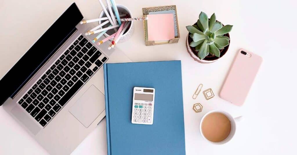 Ein Arbeitsplatz mit einem Laptop, einem blauen Notizbuch, einem Taschenrechner, Bleistiften, Büroklammern, einem Topf mit Sukkulenten, einem Smartphone und einer Tasse Kaffee.