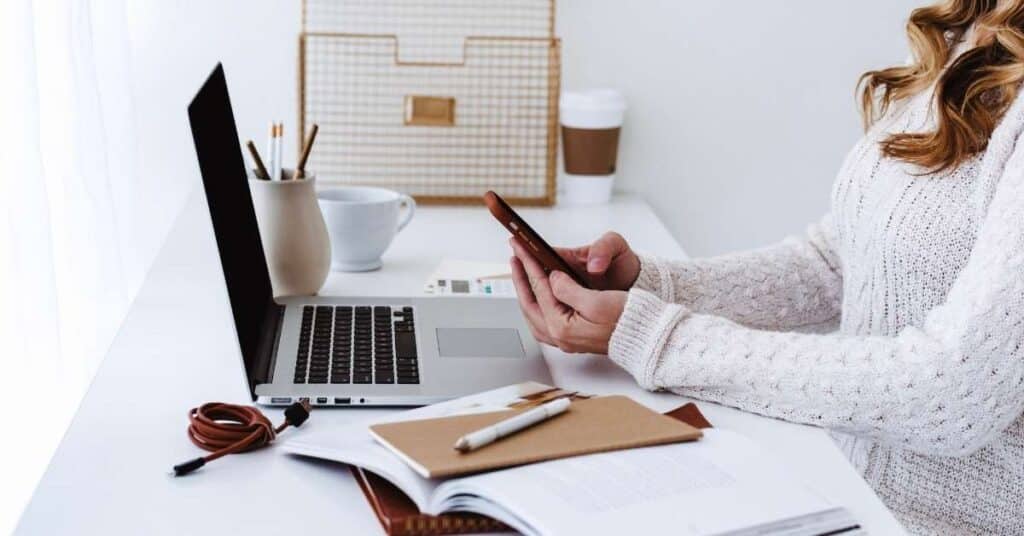 Eine Person im Pullover benutzt ein Smartphone an einem weißen Schreibtisch. Im Hintergrund sind ein Laptop, Notizbücher, Stifte, eine Kaffeetasse und eine gewebte Tasche zu sehen.