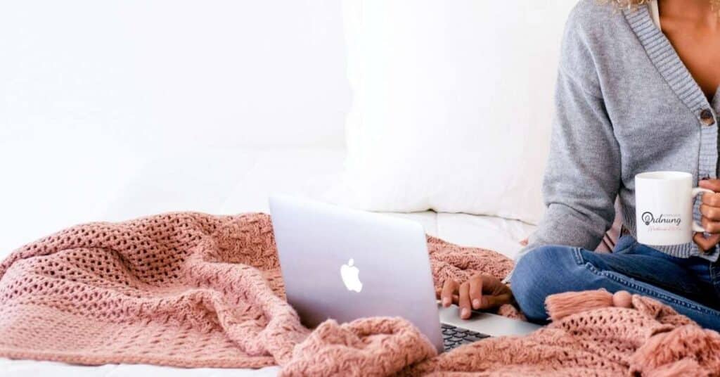Person, die mit einer rosa Decke auf einem Bett sitzt, einen Laptop benutzt und eine Tasse hält.