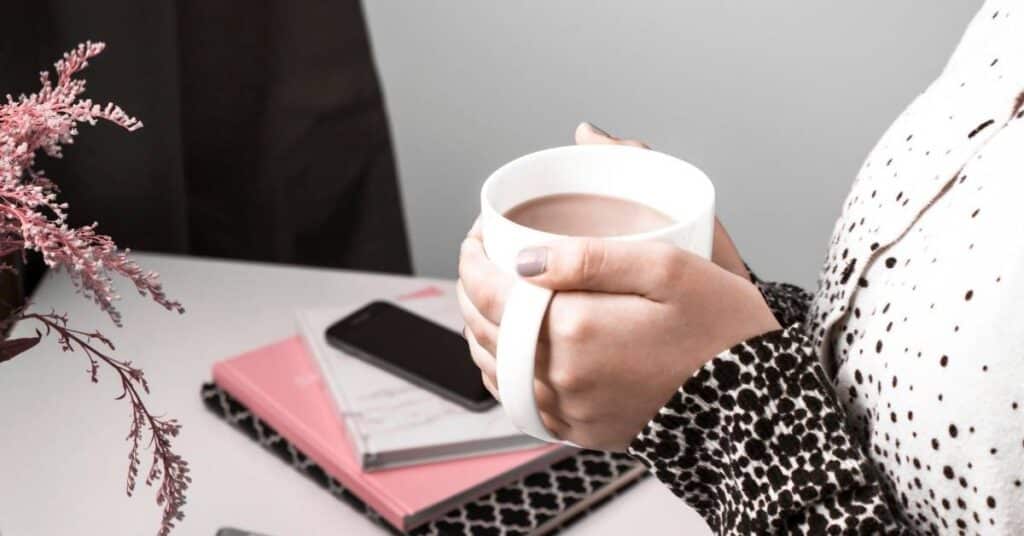 Eine Person hält einen weißen Becher neben einem Tisch mit einem Smartphone, rosa Notizbüchern und einer Vase mit Blumen.