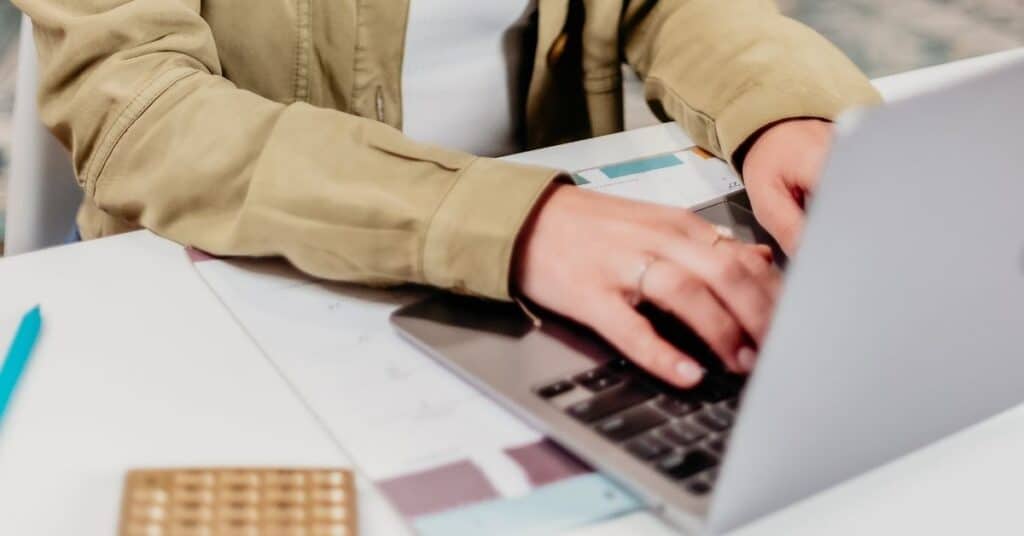 Person, die an einem Schreibtisch auf einem Laptop tippt, in dessen Nähe Papiere und ein Stift liegen.