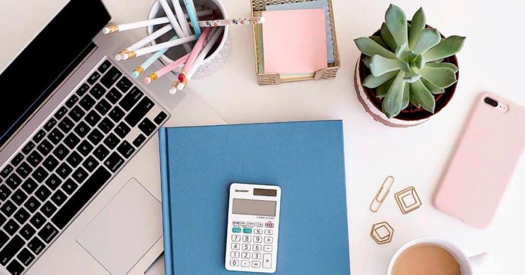 Arbeitsplatz mit Laptop, Taschenrechner auf einem blauen Ordner, Sukkulente, Smartphone, Kaffeetasse und Schreibwaren auf einem Schreibtisch.