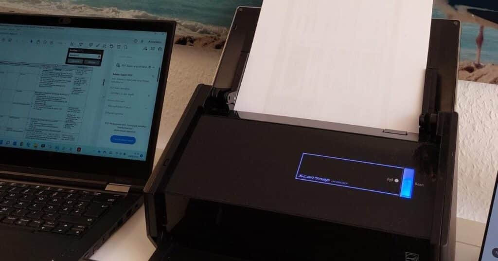 Ein Dokumentenscanner verarbeitet ein Blatt Papier. Neben dem Scanner steht ein Laptop, auf dem ein PDF-Dokument angezeigt wird.