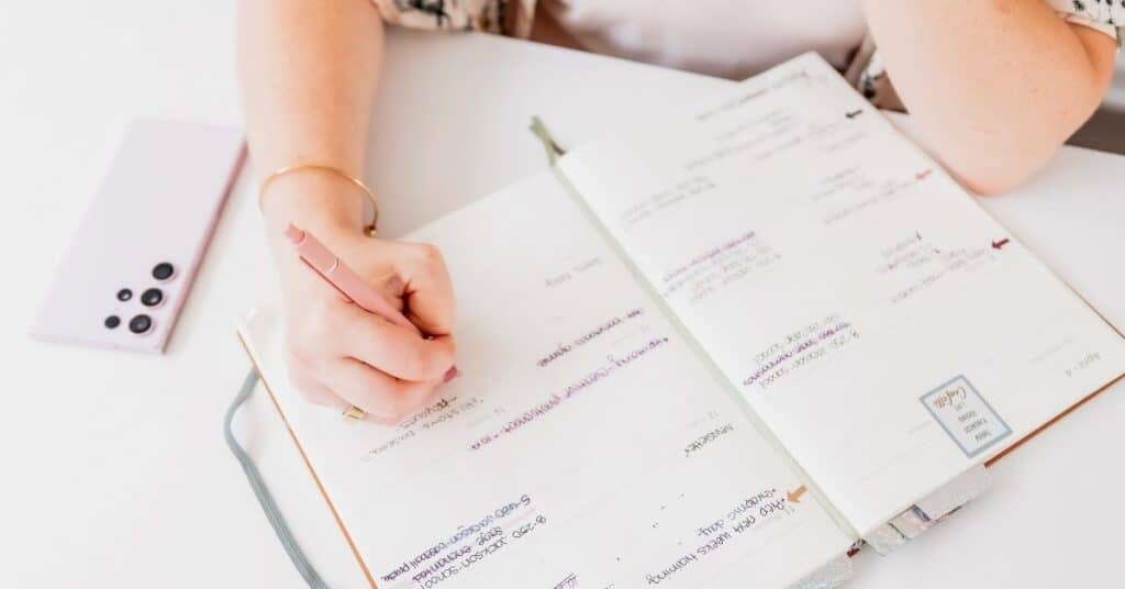 Person, die mit einem Bleistift in einen offenen Planer voller Notizen und Pläne schreibt. Ein Smartphone liegt auf dem Schreibtisch in der Nähe.