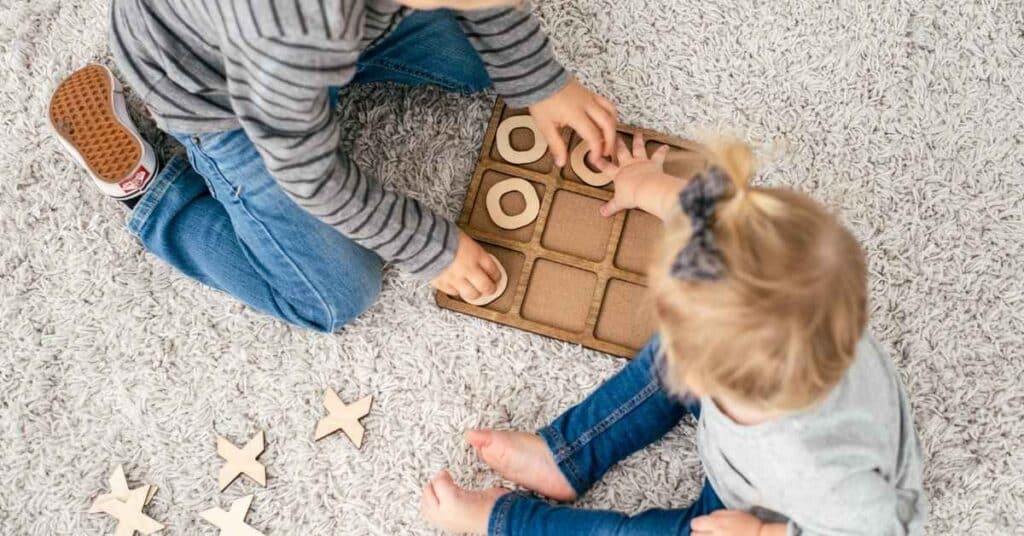 Zwei Kinder sitzen auf einem Teppich und spielen Tic-Tac-Toe mit Holzfiguren, wobei sie ihren Blick auf das Spielbrett richten.