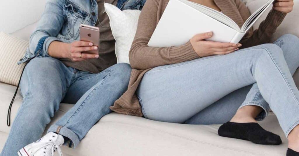 Zwei Personen sitzen auf einer Couch, eine hält ein Smartphone und die andere liest ein Buch. Beide tragen legere Kleidung, darunter Jeans und Turnschuhe.