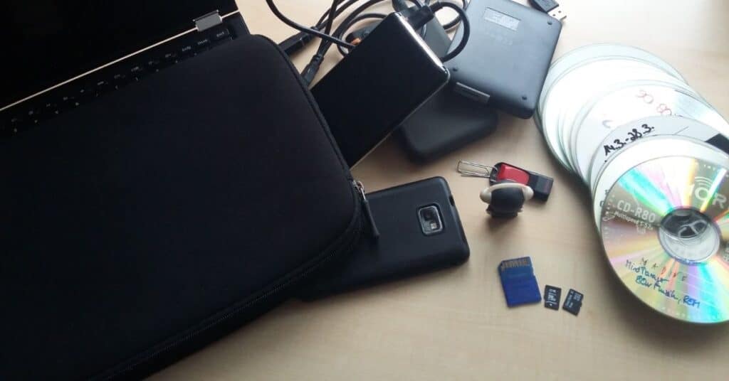 Eine Laptoptasche, externe Festplatten, ein Smartphone, USB-Sticks, SD-Karten und beschriftete CDs liegen ausgebreitet auf einer hellen Holzfläche.