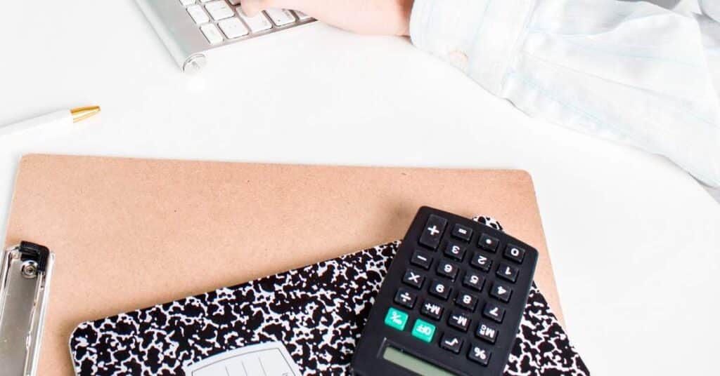 Person tippt auf einer Tastatur neben einem Klemmbrett, einem Notizbuch und einem Taschenrechner auf einem Schreibtisch.