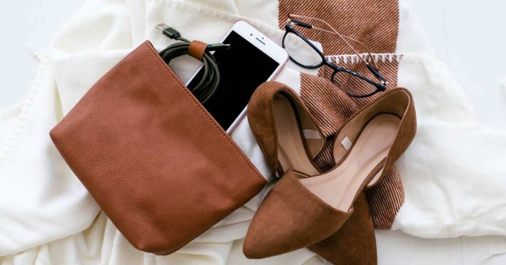 Draufsicht auf eine braune Handtasche, ein Smartphone mit Ladegerät, braune Schuhe, eine Brille und einen weiß-braunen Schal, ordentlich angeordnet.