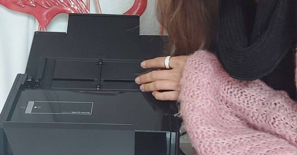 Eine Person mit einem rosa Pullover und einem schwarzen Schal legt ihre Hand auf einen geschlossenen Druckerdeckel.