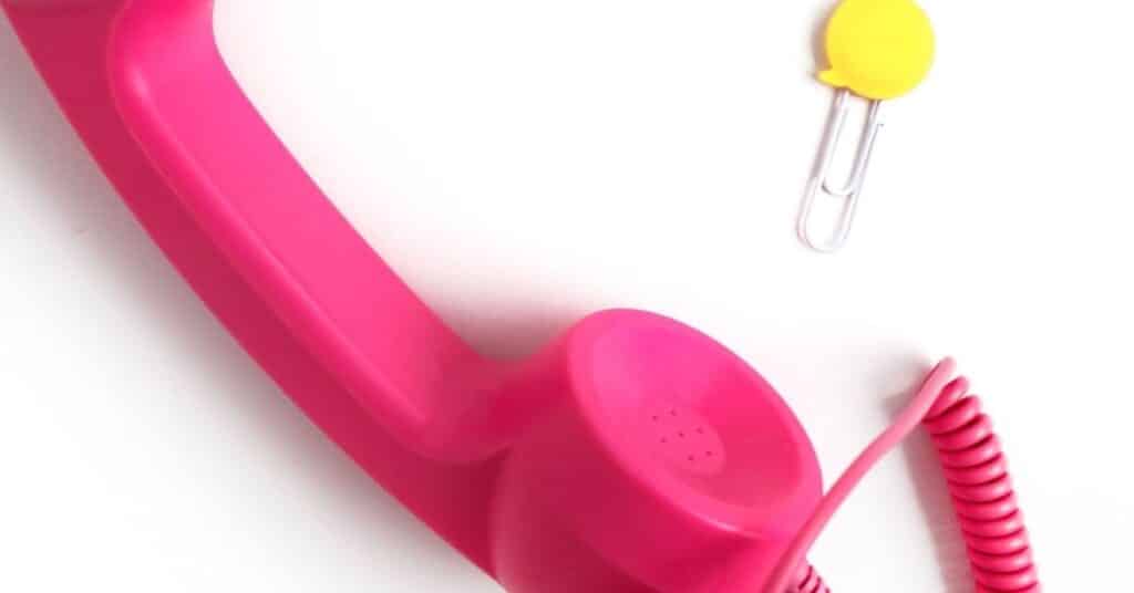 Ein rosafarbener Retro-Telefonhörer mit Spiralkabel liegt diagonal auf einer weißen Fläche neben einer Büroklammer mit einer gelben, runden Spitze.