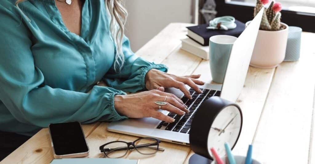 Eine Person in einer blaugrünen Bluse tippt an einem Laptop an einem Holzschreibtisch. In der Nähe stehen ein Telefon, eine Brille, eine Uhr, Tassen und Topfpflanzen.