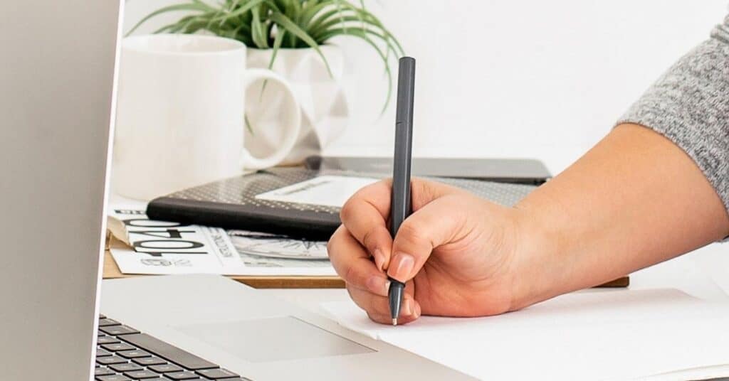 Person, die mit einem Stift auf einem Notizblock neben einem Laptop schreibt. Im Hintergrund sind eine Pflanze, eine Tasse und ein Stapel Papier zu sehen.