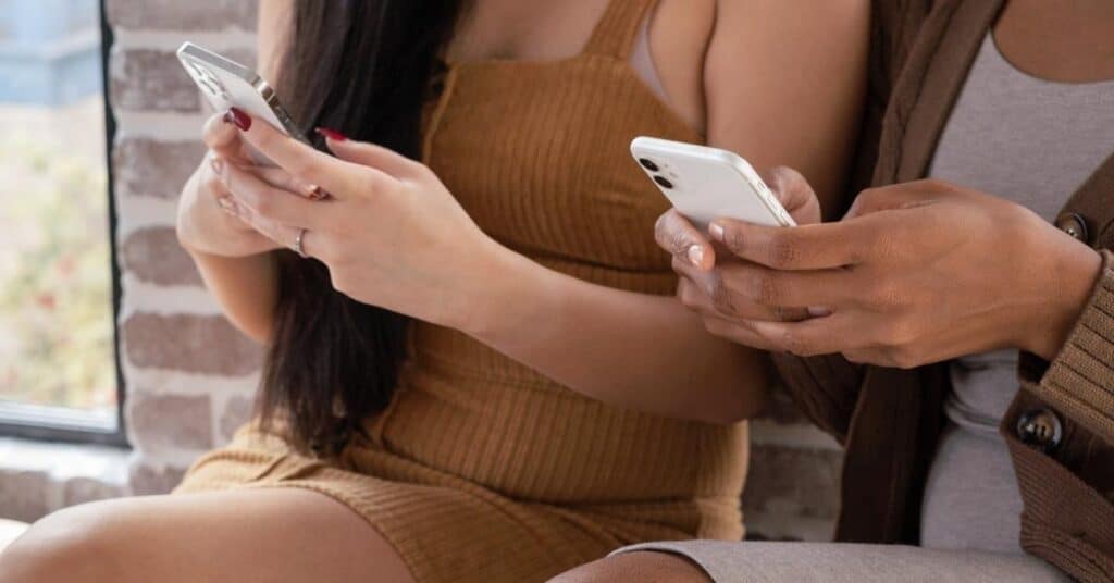 Zwei Personen sitzen nebeneinander und benutzen Smartphones. Eine trägt ein braunes Kleid und die andere eine braune Jacke. Im Hintergrund ist eine Backsteinmauer zu sehen.