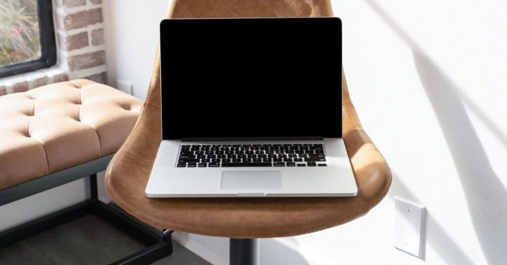 Ein geschlossener Laptop auf einem braunen Stuhl neben einem Fenster mit einer Ziegelwand und einer beige gepolsterten Bank.