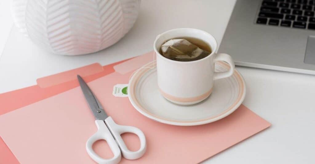 Eine Tasse Tee auf einer Untertasse steht neben einer Schere und rosa Ordnern auf einem Schreibtisch, auf dem teilweise ein Laptop zu sehen ist.